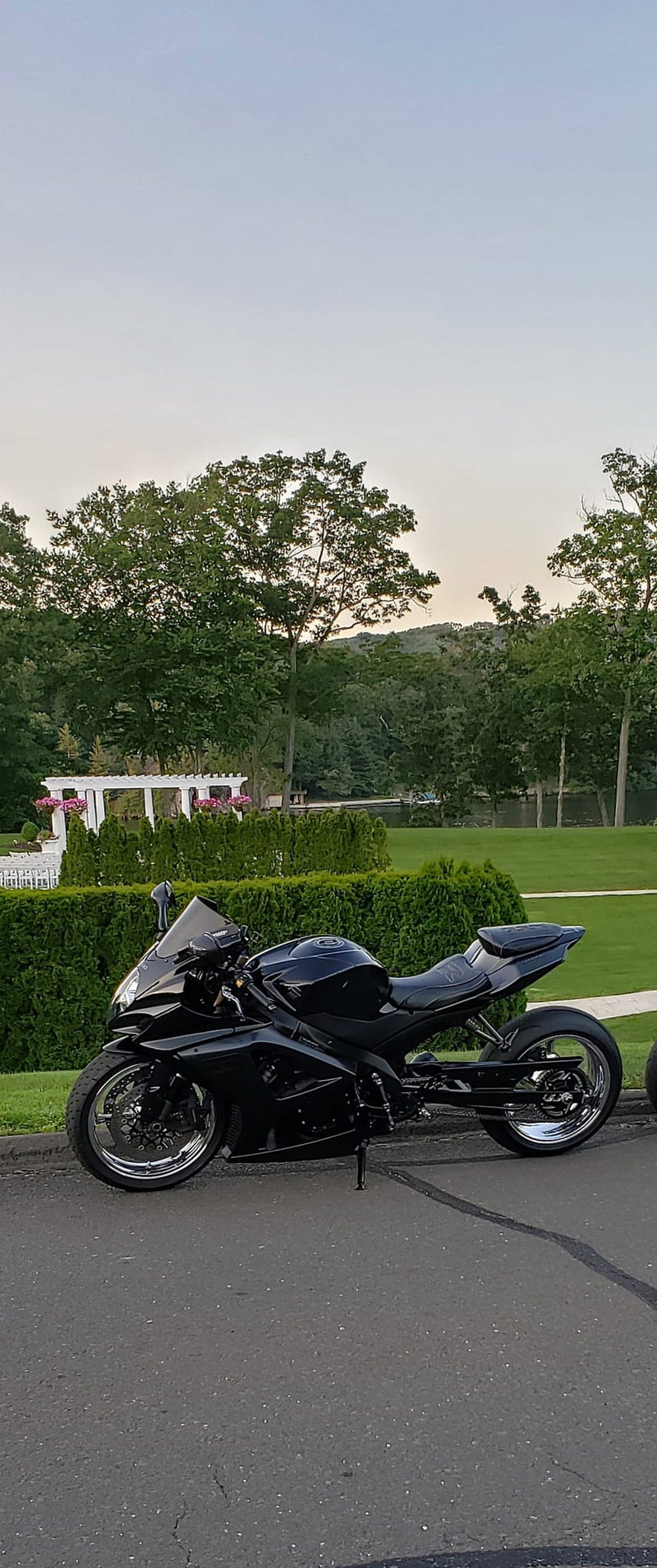Black Suzuki sportbike parked on asphalt near a white pergola with pink flowers and lush green garden hedges.