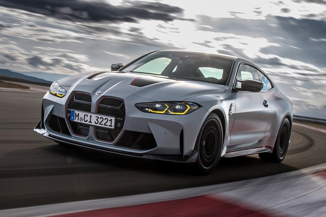Silver BMW M4 CSL with yellow headlights and red accents driving fast on a racetrack under a cloudy sky.