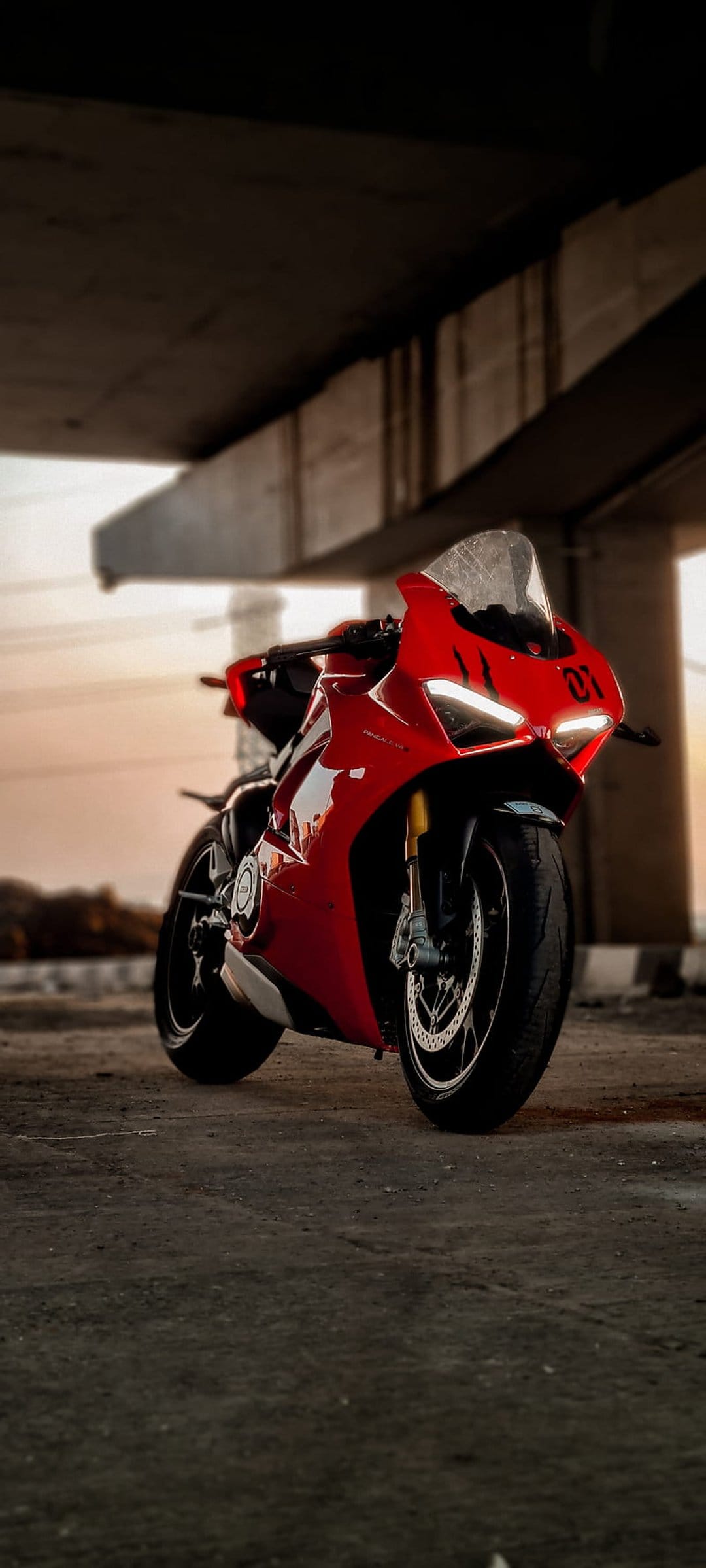 Red Ducati motorcycle with glowing headlights parked under a concrete bridge during a soft sunset at dusk.