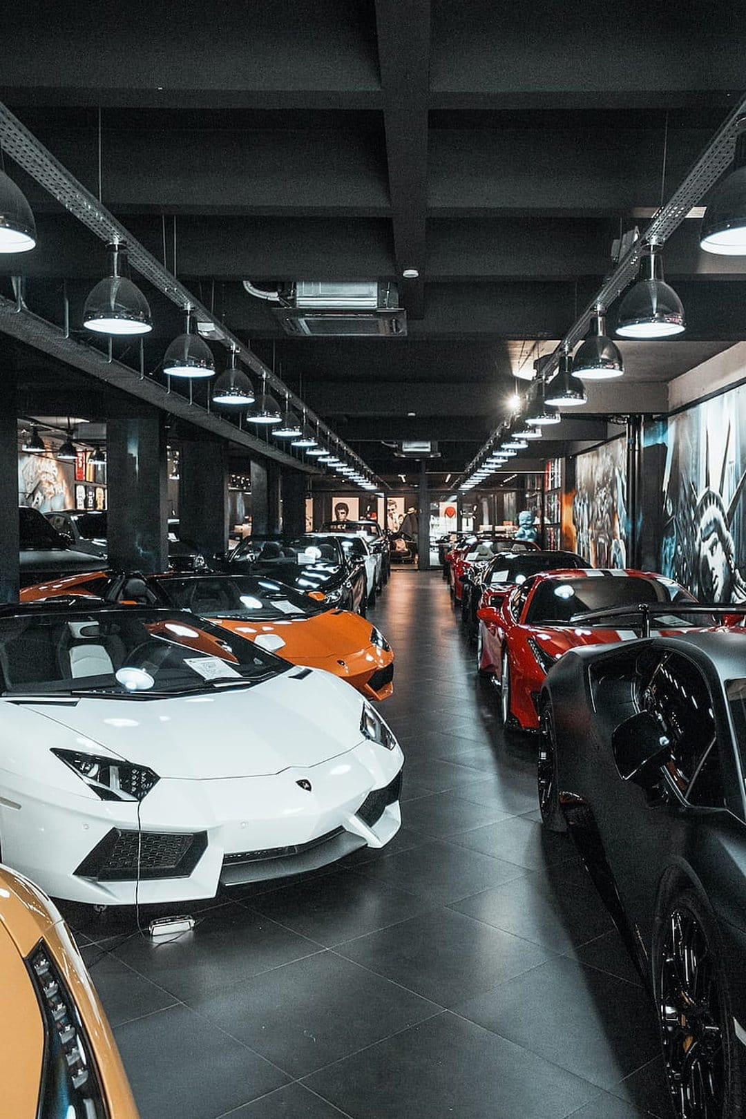 White, orange, and red supercars parked on a glossy black showroom floor under hanging industrial lamps.