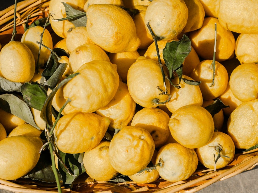 Yellow lemons with green leaves fill a brown woven basket in a top-down view for a refreshing mobile display.