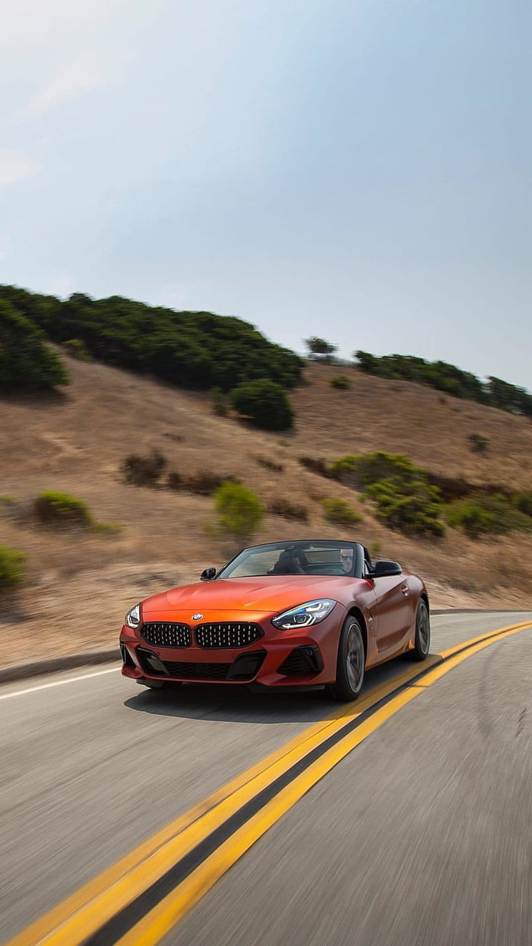Orange BMW Z4 convertible speeds along a winding asphalt road through lush green hills under soft light.