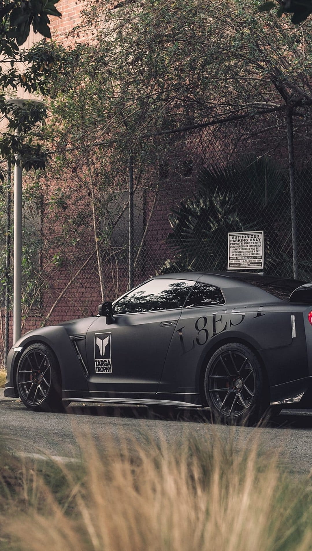 Matte black Nissan GT-R with Targa Trophy decals parked by a brick wall and tall grass in a vertical composition.