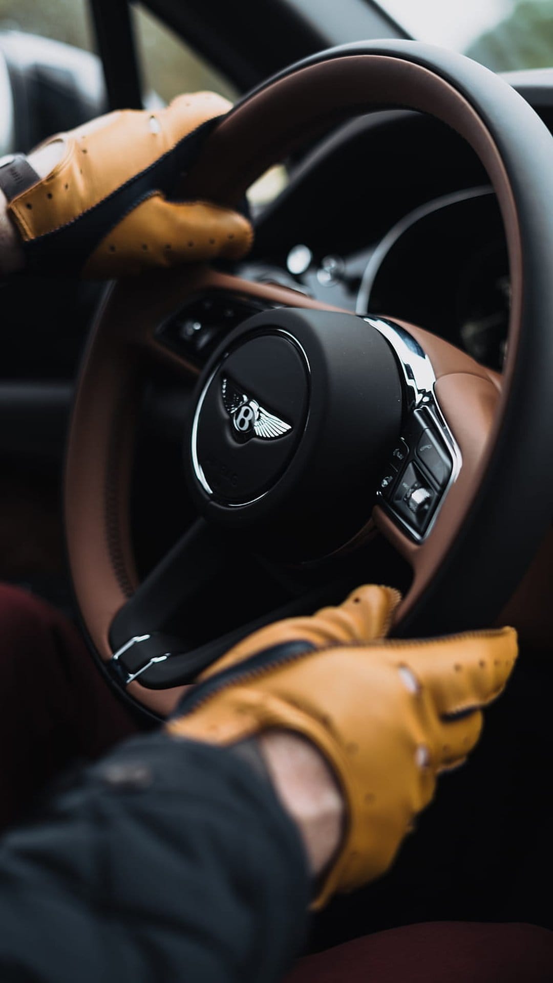 Tan leather gloves grip a Bentley steering wheel with a silver emblem against a rich brown cabin interior.