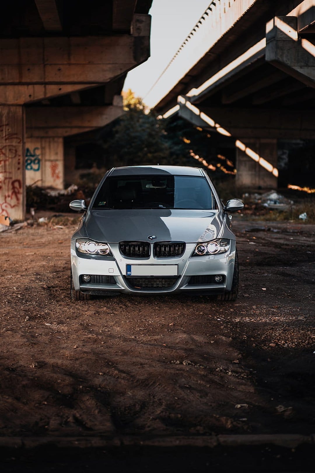 Silver BMW sedan parked on dirt ground beneath a concrete bridge with graffiti and dappled sunlight.