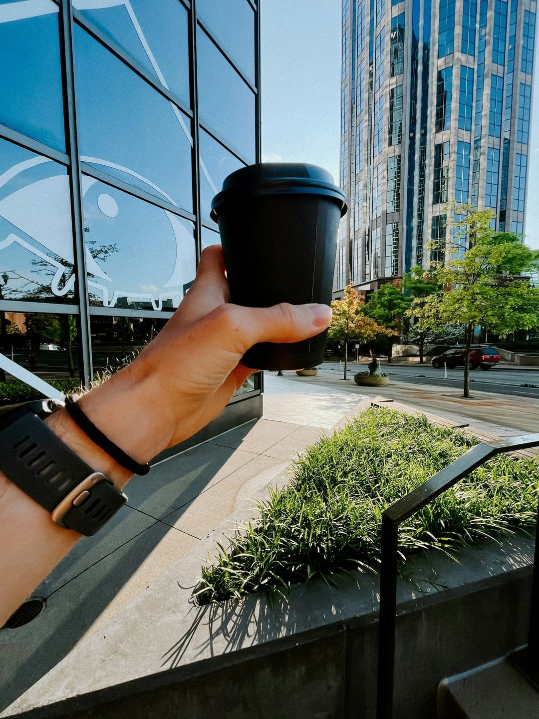 Hand holding a black disposable coffee cup against a backdrop of glass architecture and green foliage.