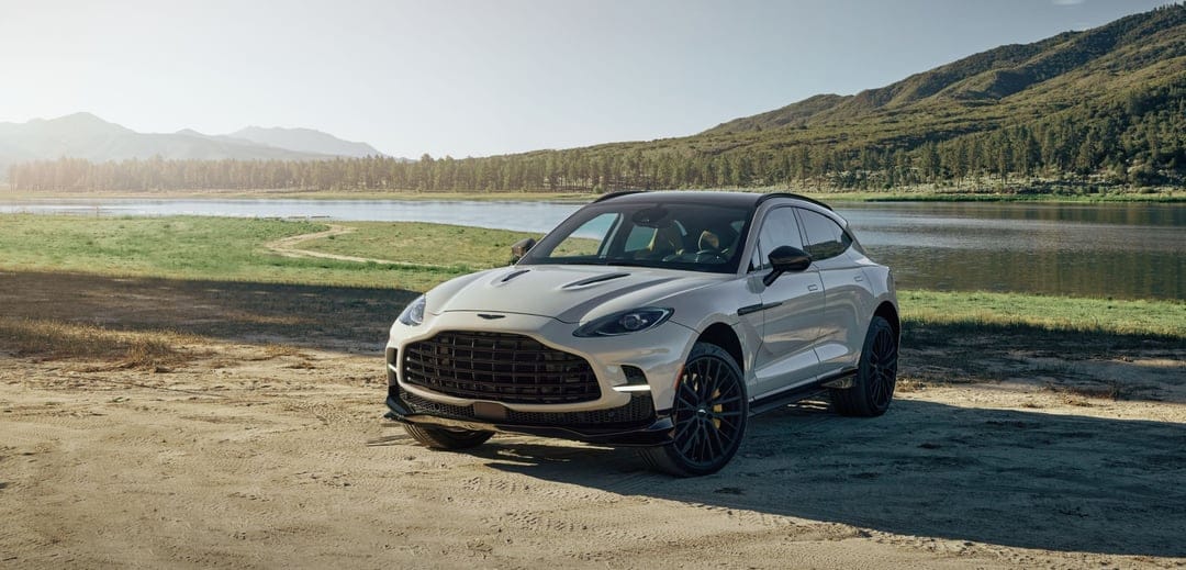 White Aston Martin DBX707 parked on a sandy shore near a calm lake with forested mountains under a soft sun.