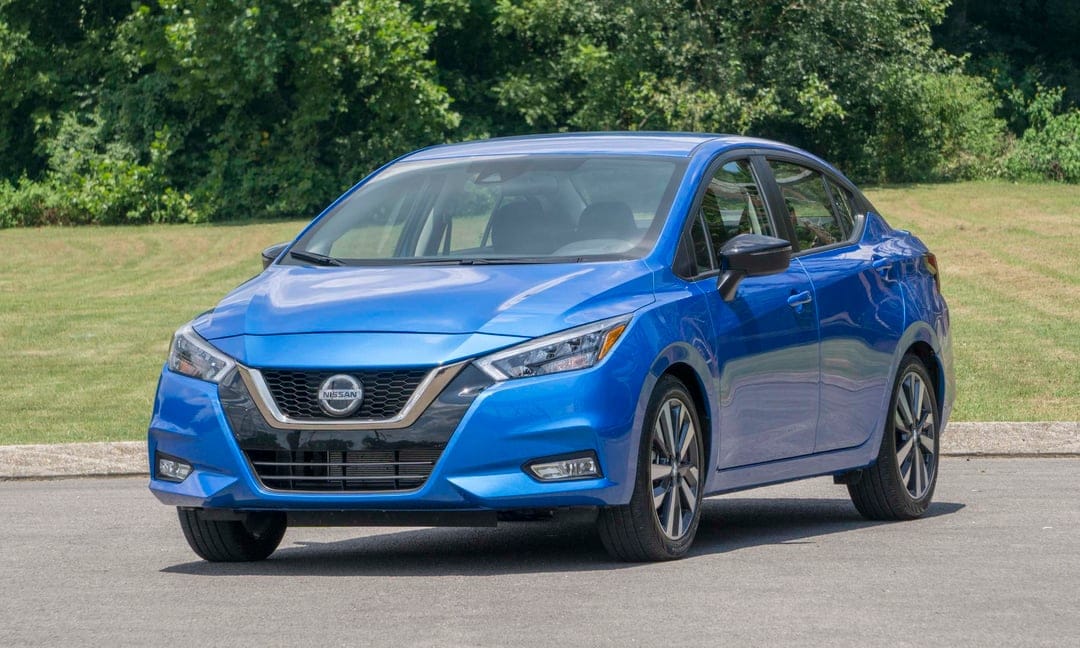 Blue Nissan sedan parked on a grey asphalt road with lush green trees and a grassy field in the background.