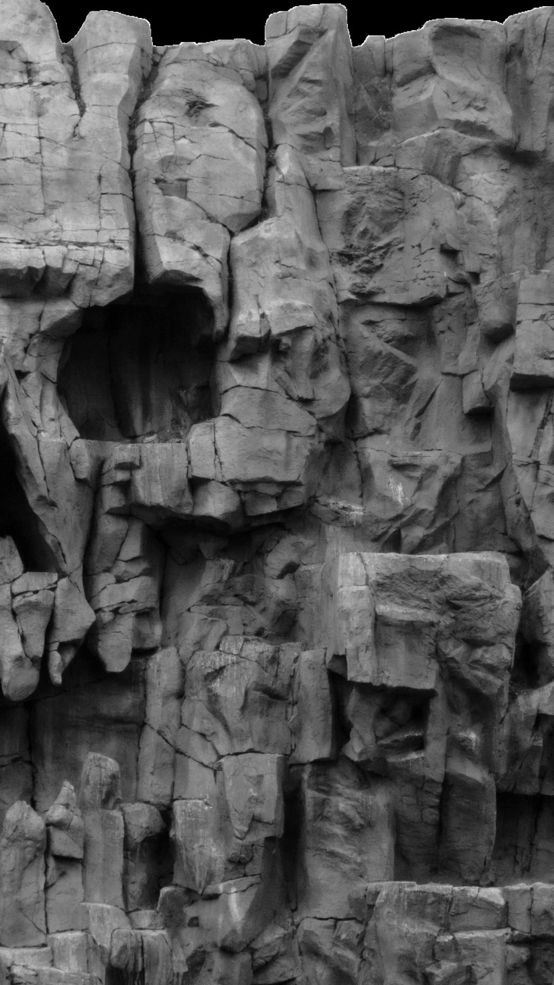 Jagged rock cliff featuring deep vertical cracks and high-contrast shadows in a monochromatic portrait view.