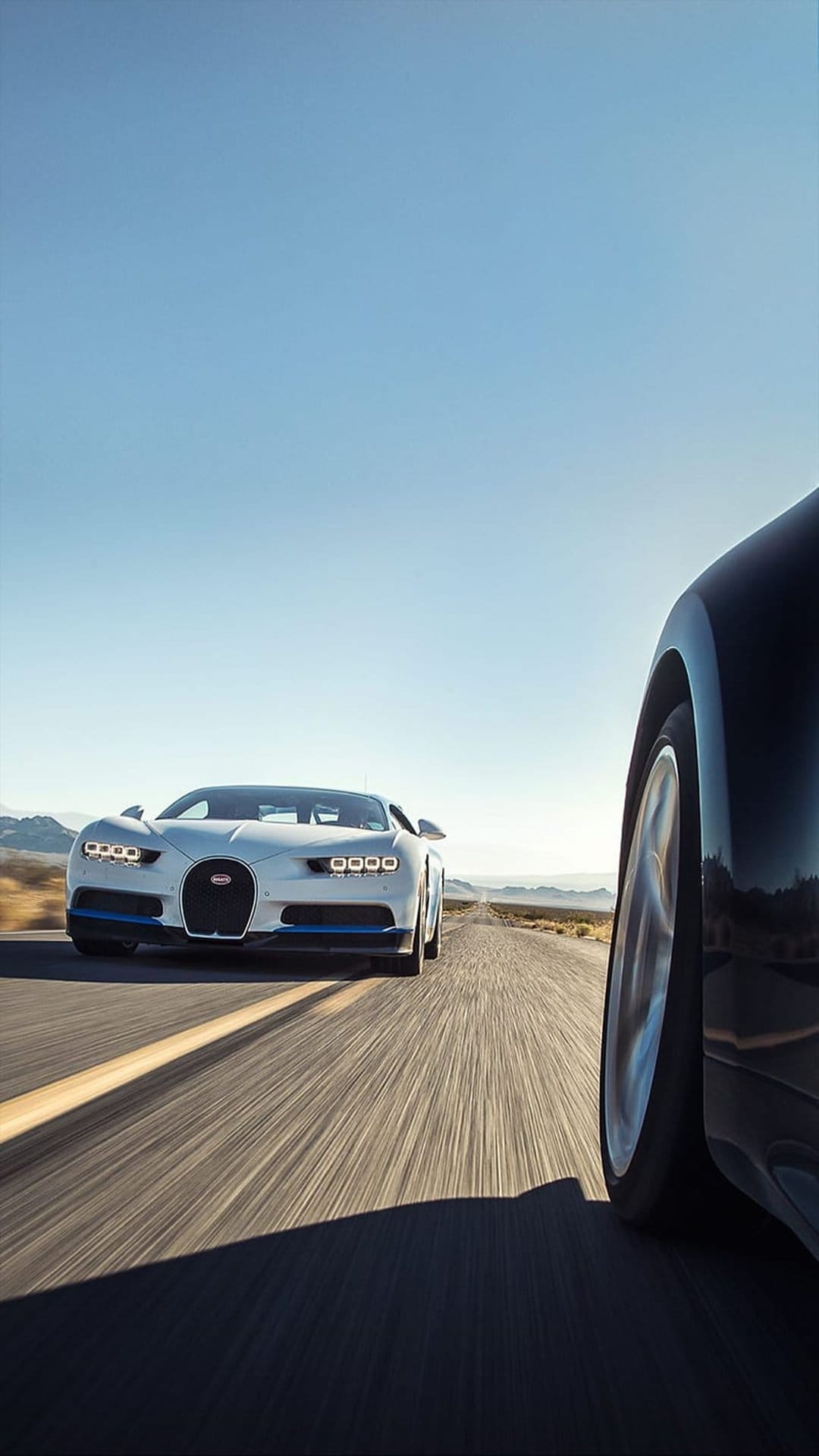 White and black Bugatti hypercars race on a sunlit desert road under a clear blue sky with motion blur.