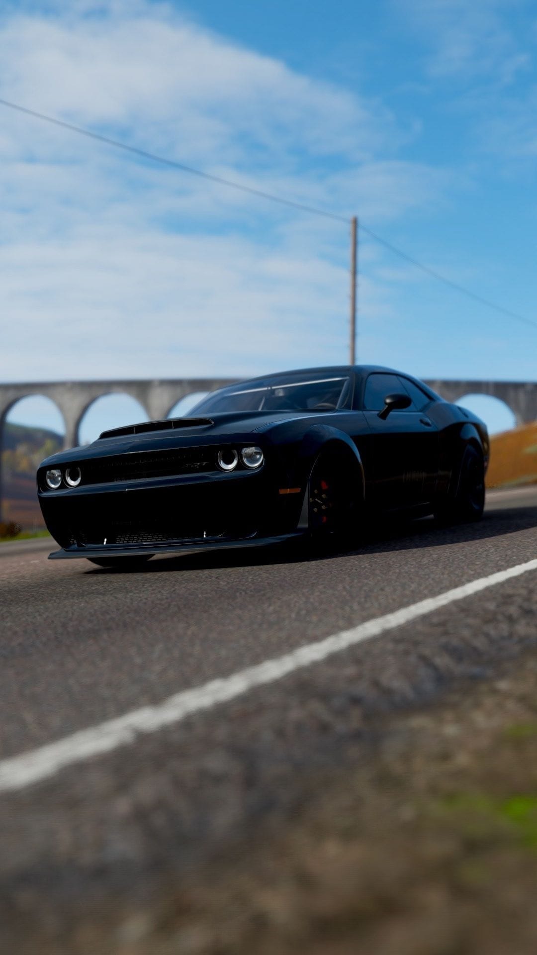 Black muscle car on a paved road with a stone arch bridge and blue sky in the background under bright daylight.