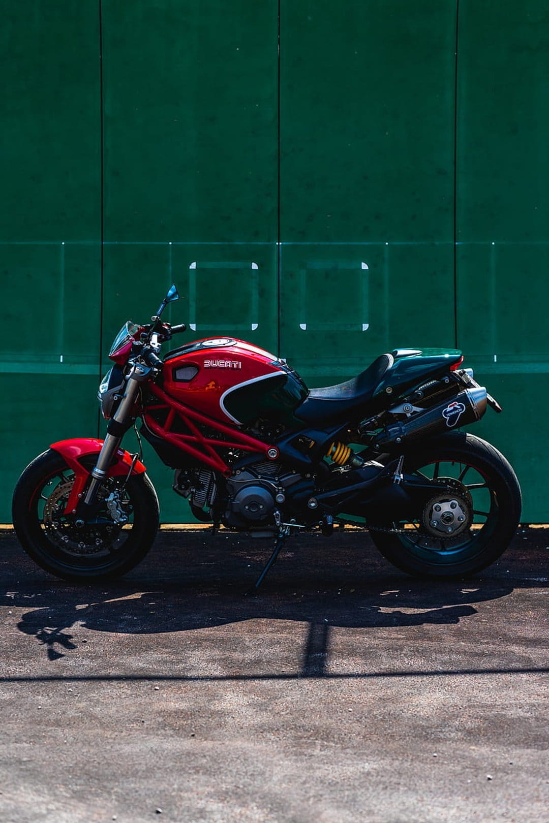 Red Ducati motorcycle with a trellis frame stands before a textured green wall on sunlit asphalt ground.