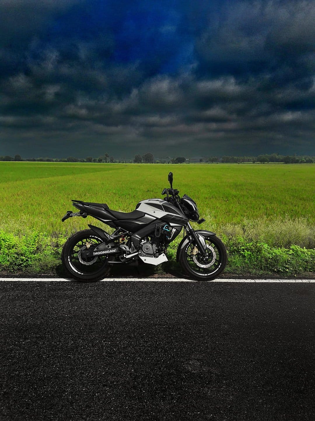 Silver motorcycle parked on an asphalt road beside a green field beneath a dark, swirling stormy sky.