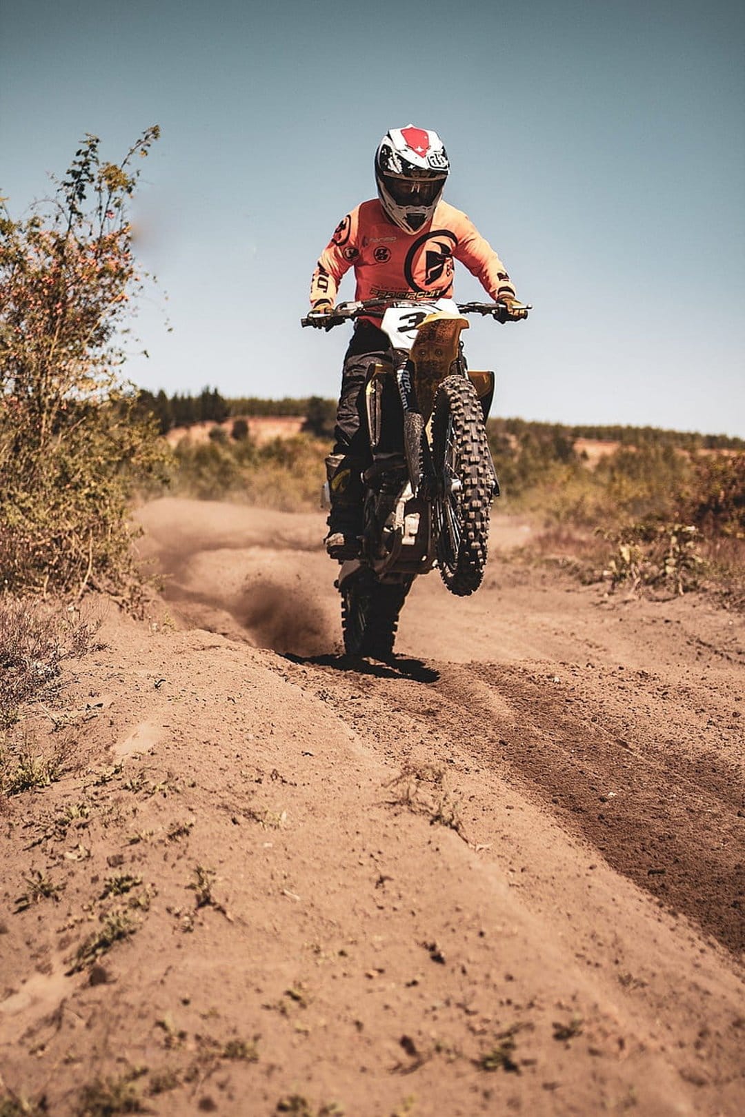 Motocross rider in orange gear performing a wheelie on a dirt track under a blue sky with rising dust clouds.
