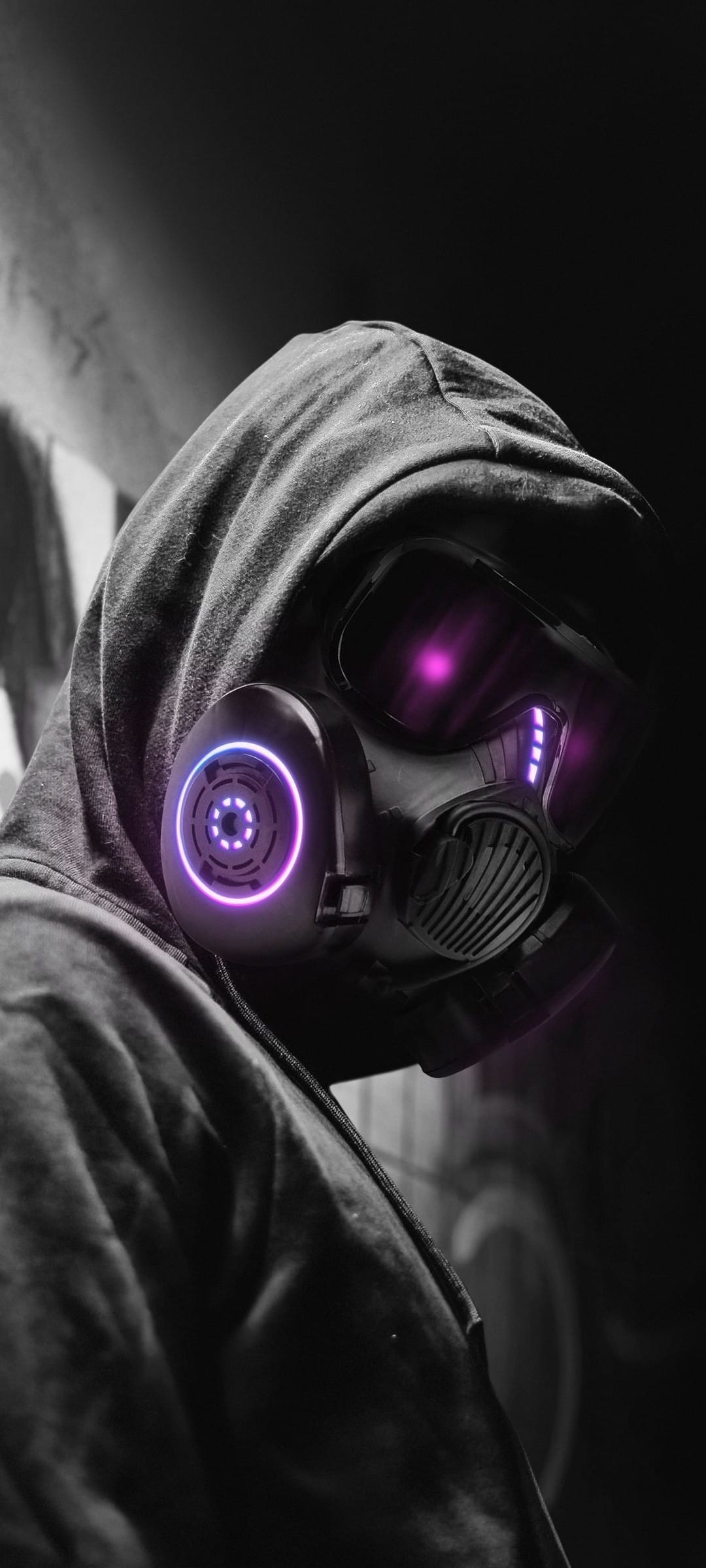 Hooded person wearing a black gas mask with glowing purple lenses and blue circular lights in a dark setting.