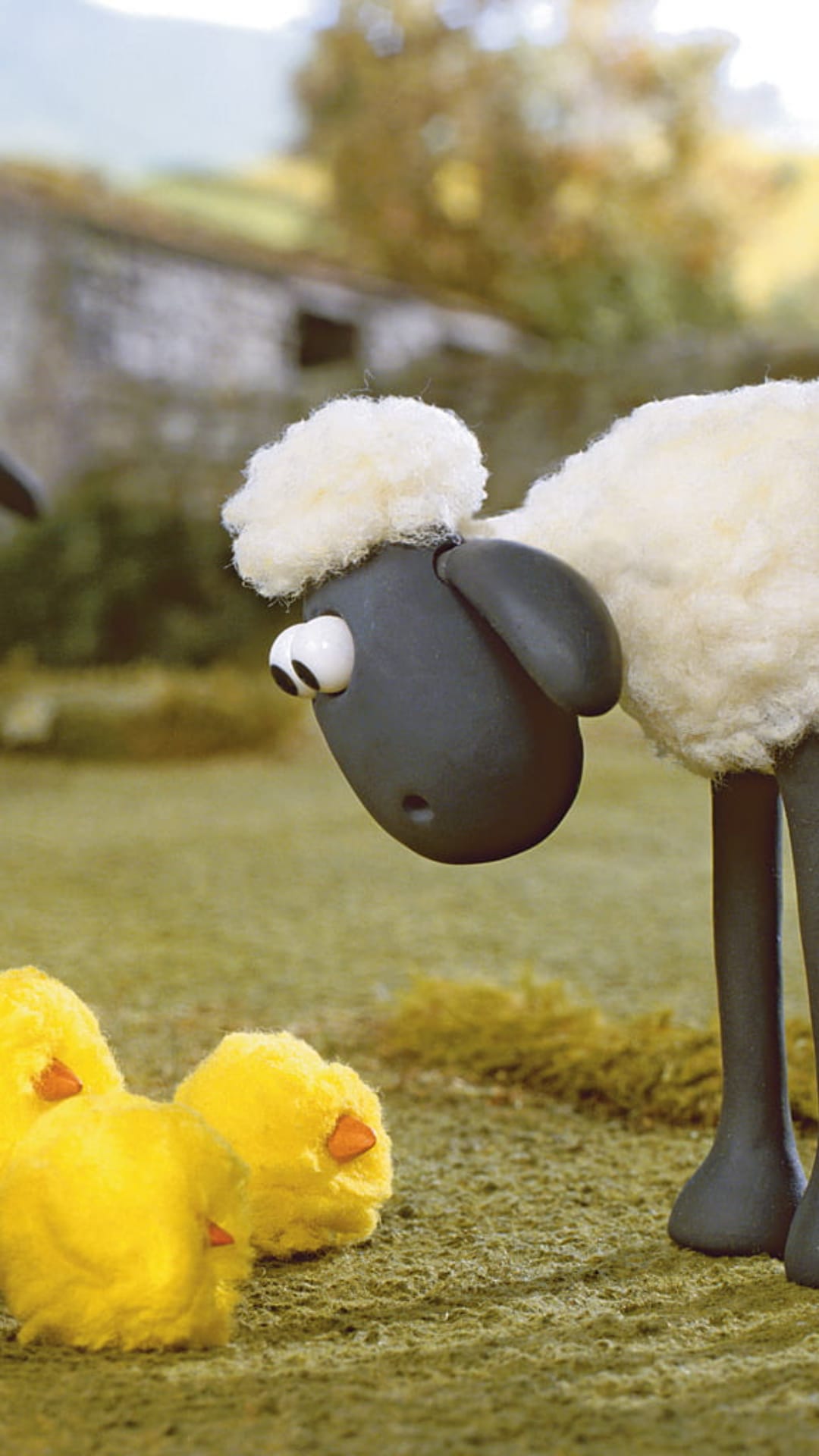 Shaun the Sheep stares down at three tiny yellow chicks on a grassy farmyard with a stone barn in the distance.