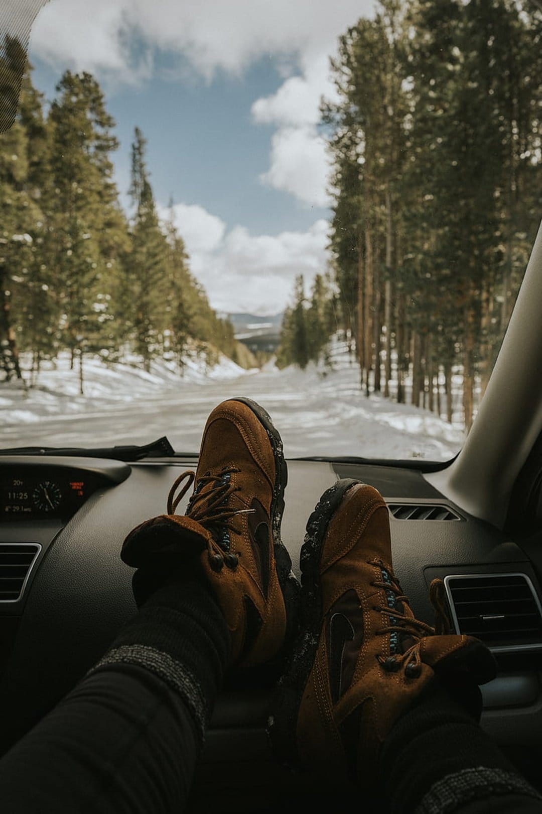 Brown leather hiking boots rest on a dashboard overlooking a snowy road lined with tall pine trees in winter.