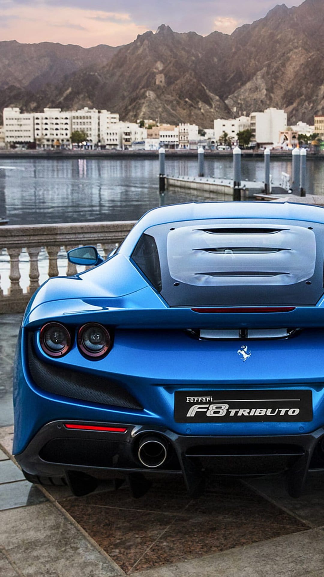 Blue Ferrari F8 Tributo rear view on a stone patio with a lake and mountains under a clear sky in the background.