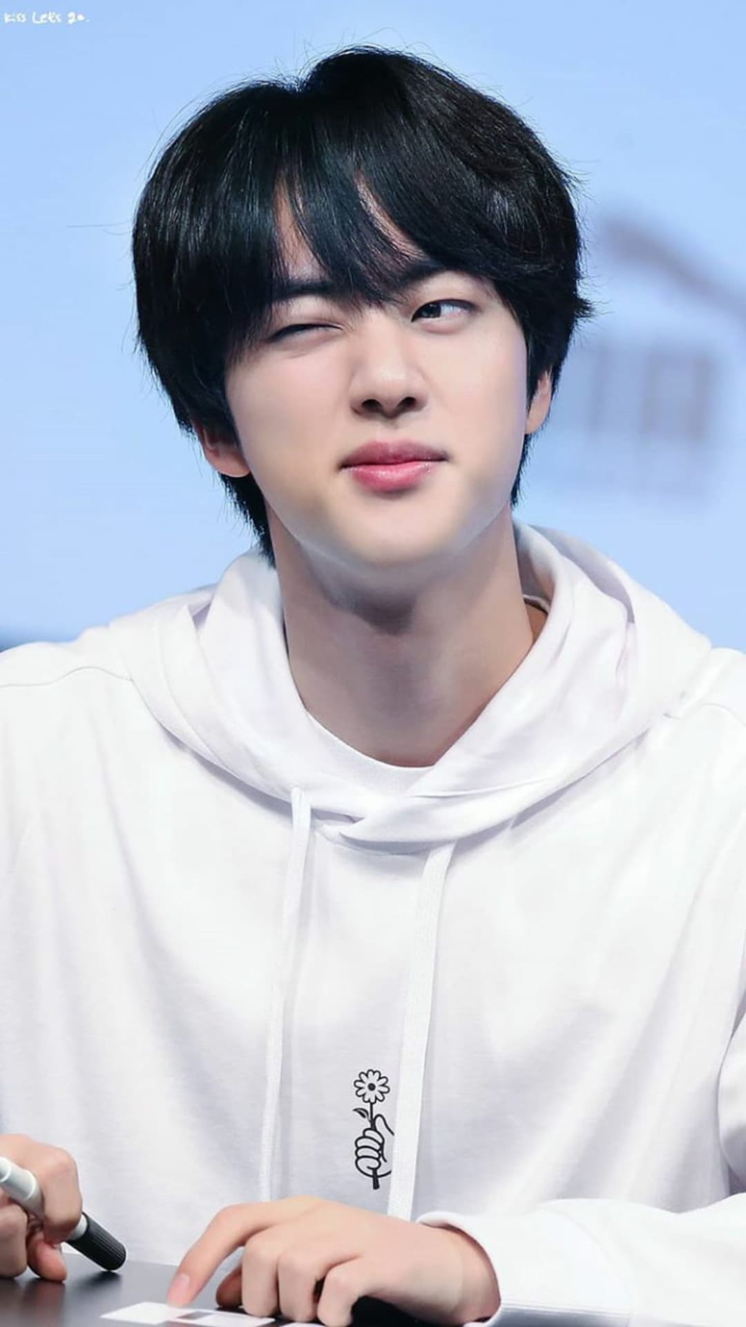Jin from BTS winks playfully while wearing a white hoodie with a flower graphic against a soft blue background.