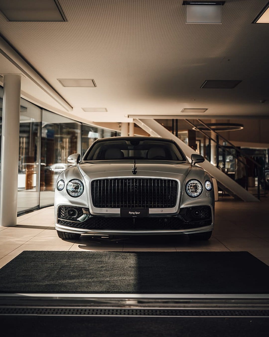 Silver Bentley Flying Spur sedan with glowing headlights parked on a polished floor in a glass-walled showroom.
