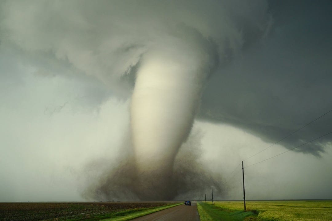 Massive dark tornado funnel touching down on a flat rural landscape with swirling debris under stormy skies.