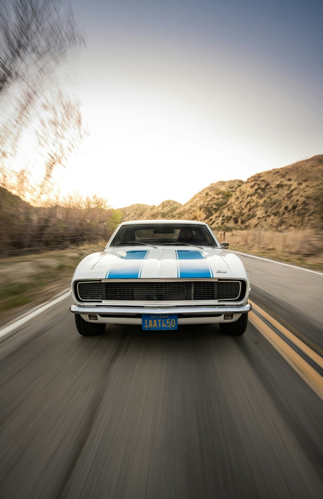 White Chevrolet Camaro with blue stripes racing on a highway at sunset with mountains in the background.