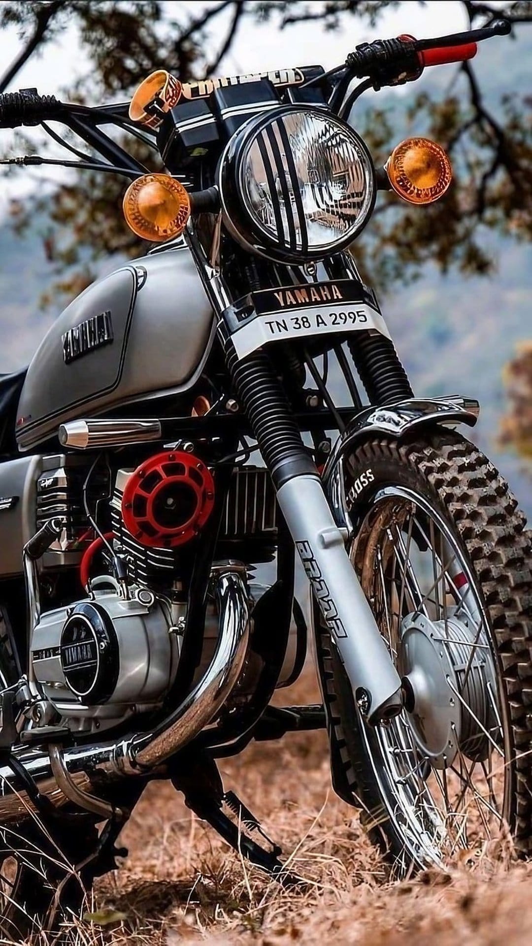 Vintage Yamaha motorcycle featuring a silver fuel tank, round caged headlight, and off-road tires on dry grass.