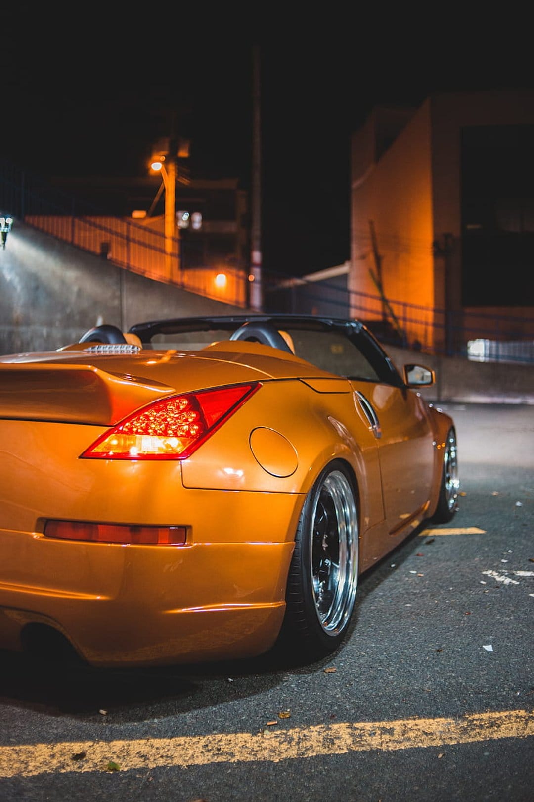 Orange convertible sports car with glowing taillights parked on a dark urban street under amber streetlights.