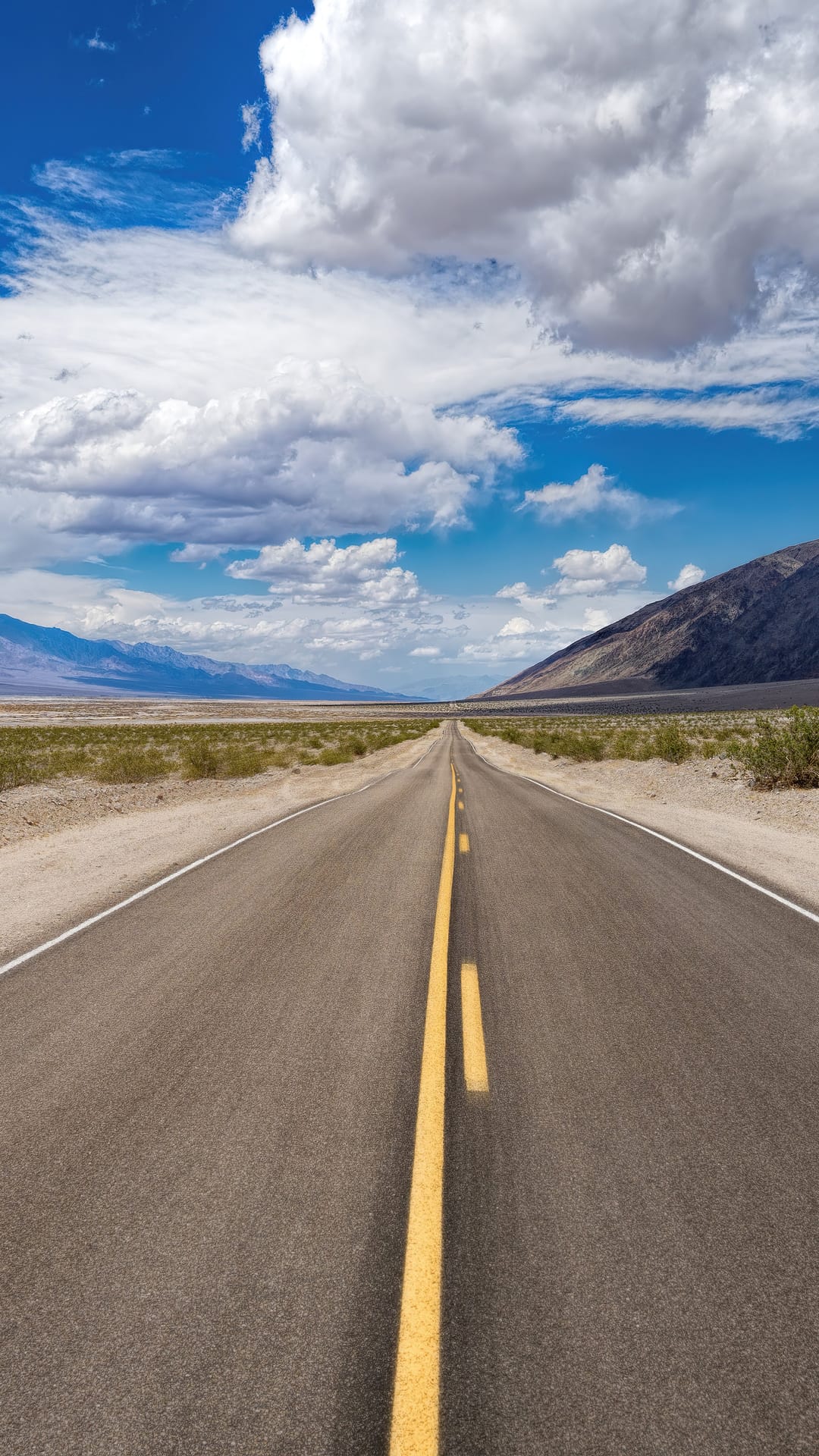 Asphalt road stretching toward hazy mountains under a bright blue sky filled with white cumulus clouds.