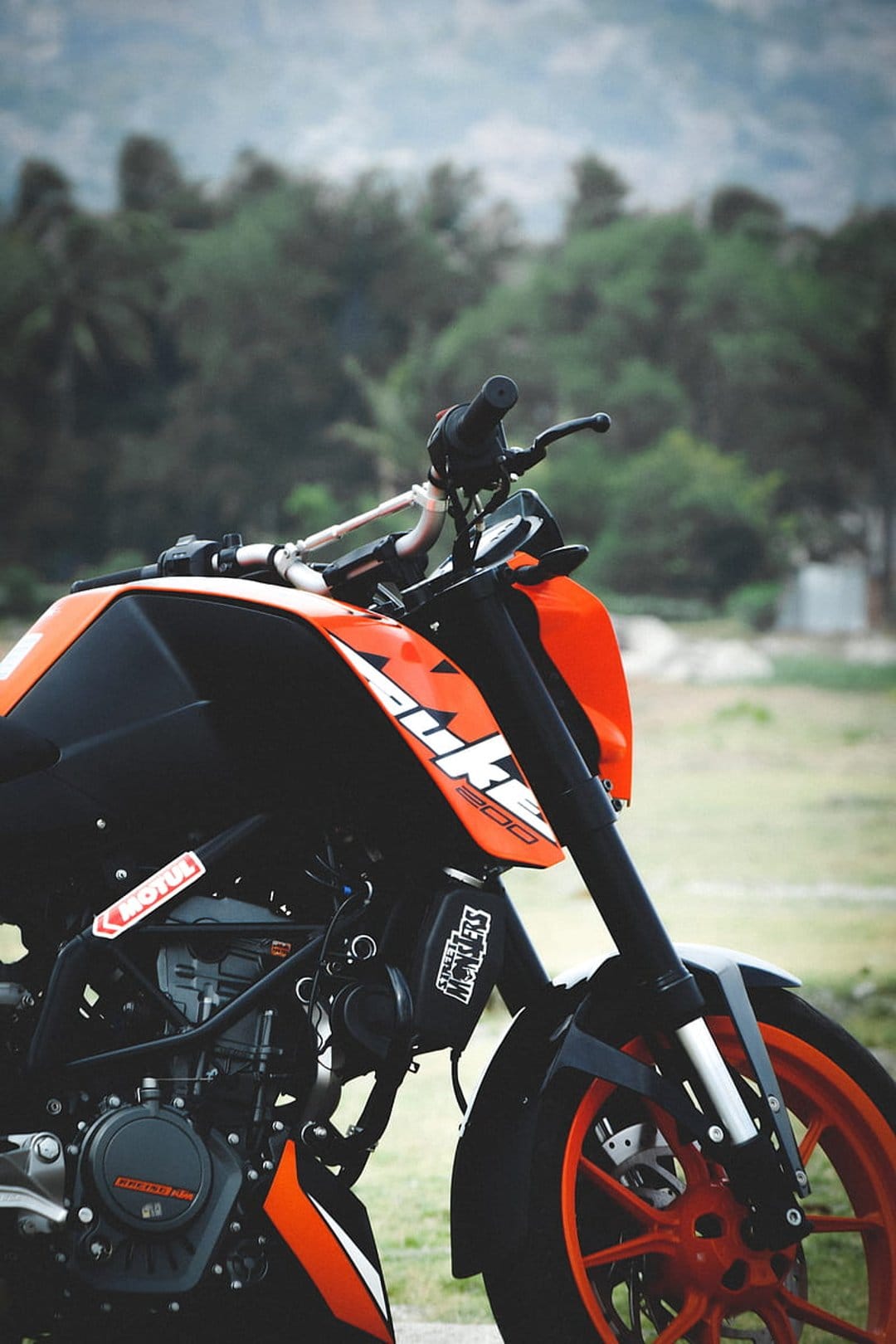 KTM Duke 200 motorcycle featuring an orange tank and black handlebars with a blurred green forest background.
