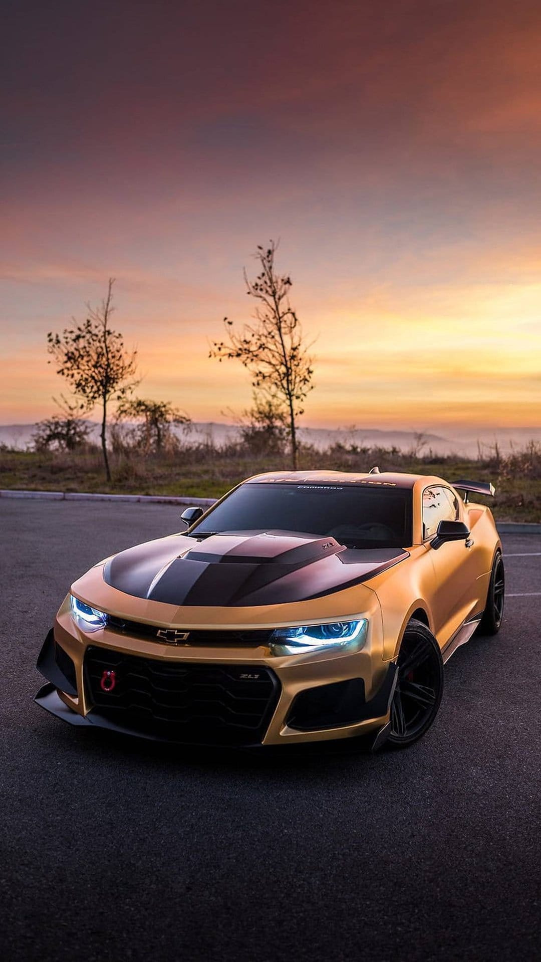 Golden Chevrolet Camaro ZL1 with racing stripes parked on asphalt against a vibrant purple and orange sunrise.