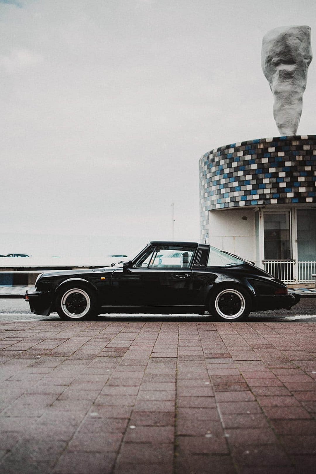 Black Porsche Targa on brick pavement near a blue mosaic building and abstract sculpture under gray skies.