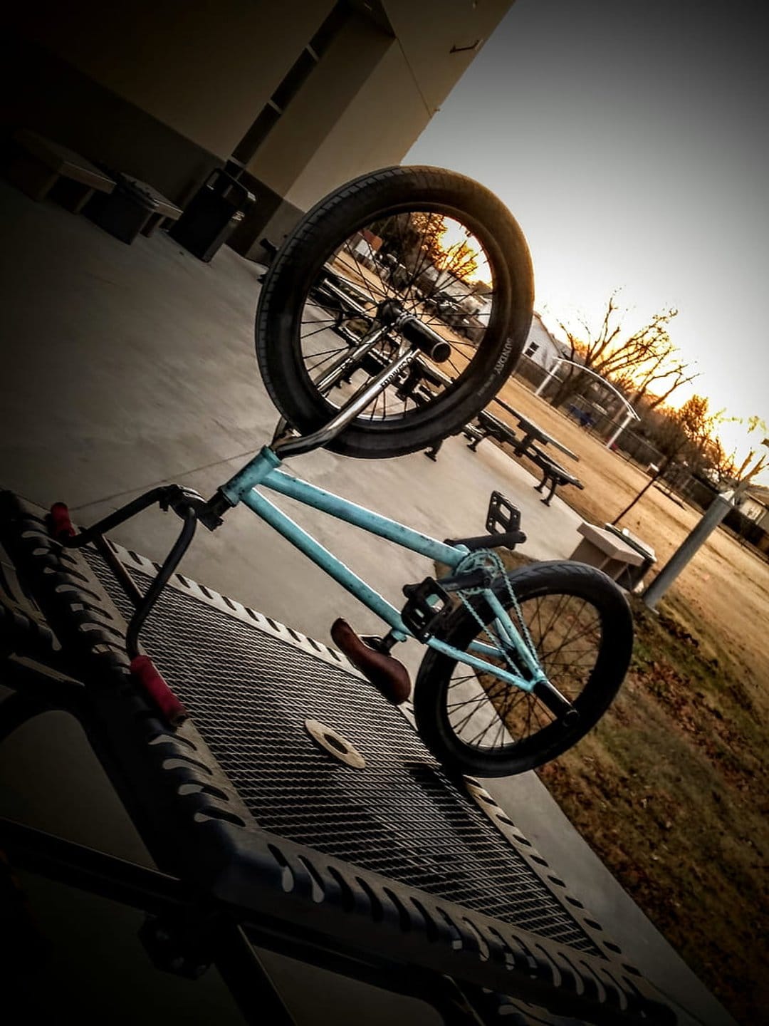 Light blue BMX bike rests at a tilt on a trampoline in a grassy park during a vibrant orange sunset.