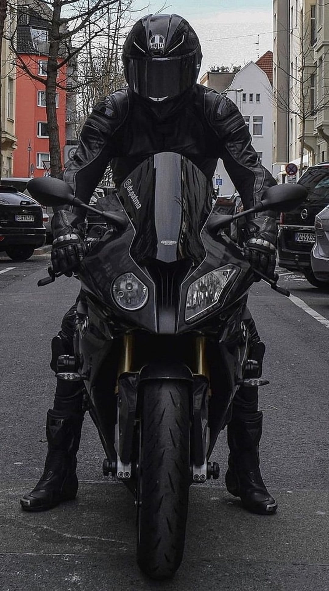 Motorcyclist in black leather gear sits on a sports bike with headlights on against a city street backdrop.