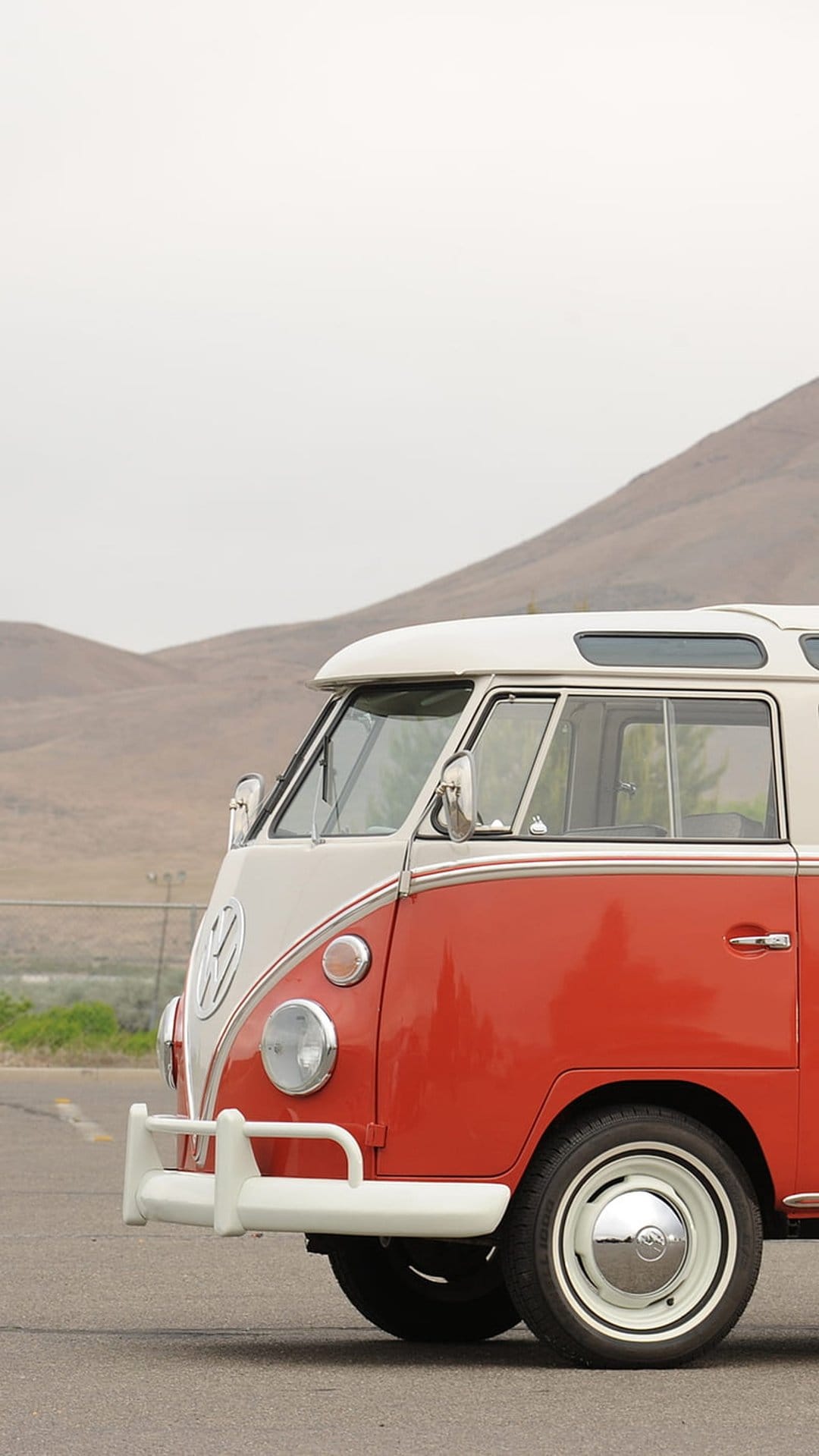 Red and white vintage VW bus front view with chrome bumper on an asphalt road with dry hills in the background.