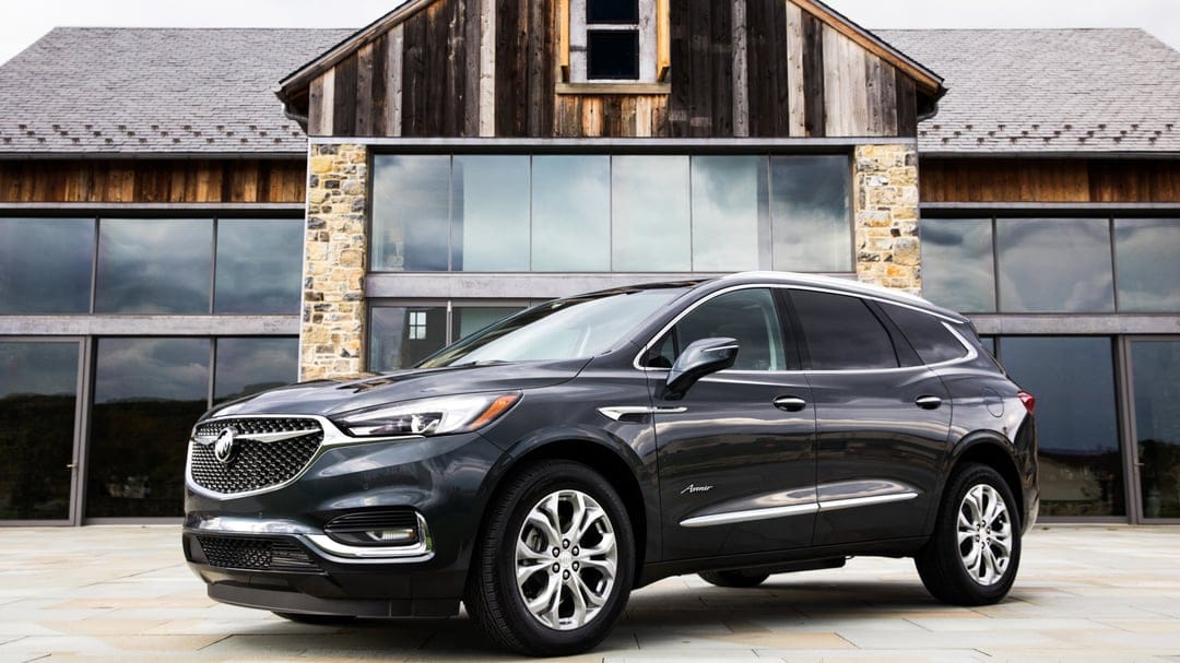 Buick Enclave Avenir SUV parked on a stone patio in front of a modern barn-style building with large glass windows.
