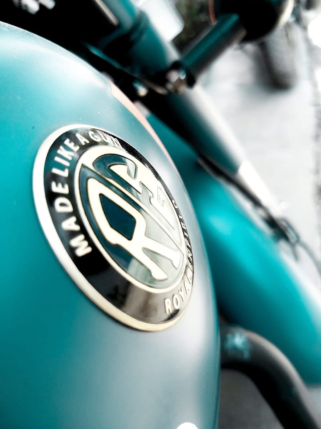 Royal Enfield emblem with Made Like A Gun text on a matte teal motorcycle fuel tank with a blurred background.