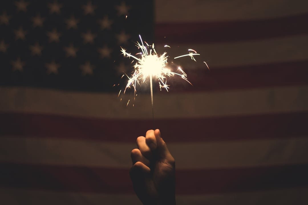 Hand-held sparkler emitting bright white light and glowing embers in front of a blurred red and white striped flag.