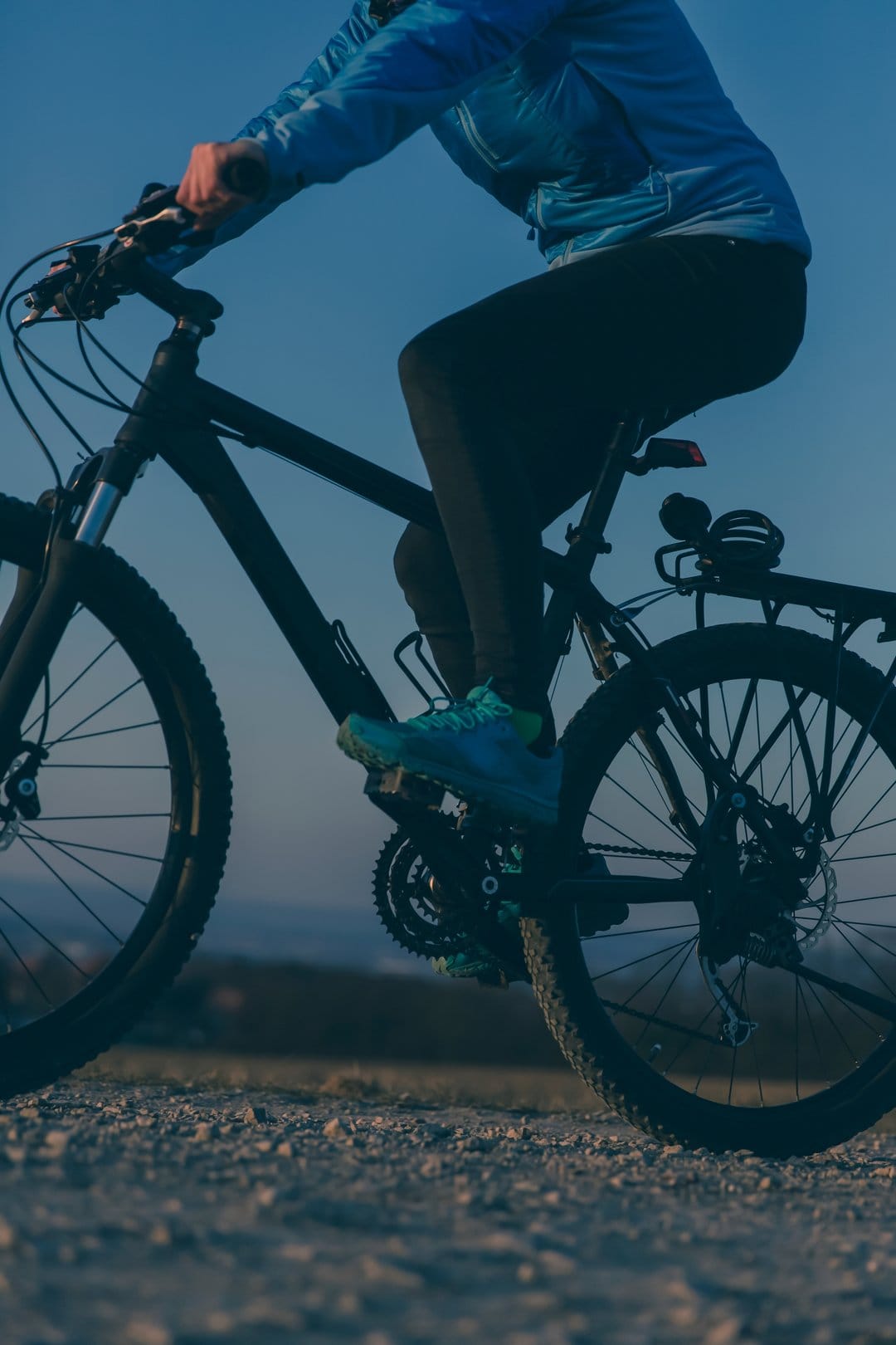 Cyclist in a blue jacket and black leggings rides a mountain bike on a sunny gravel path under a clear blue sky.