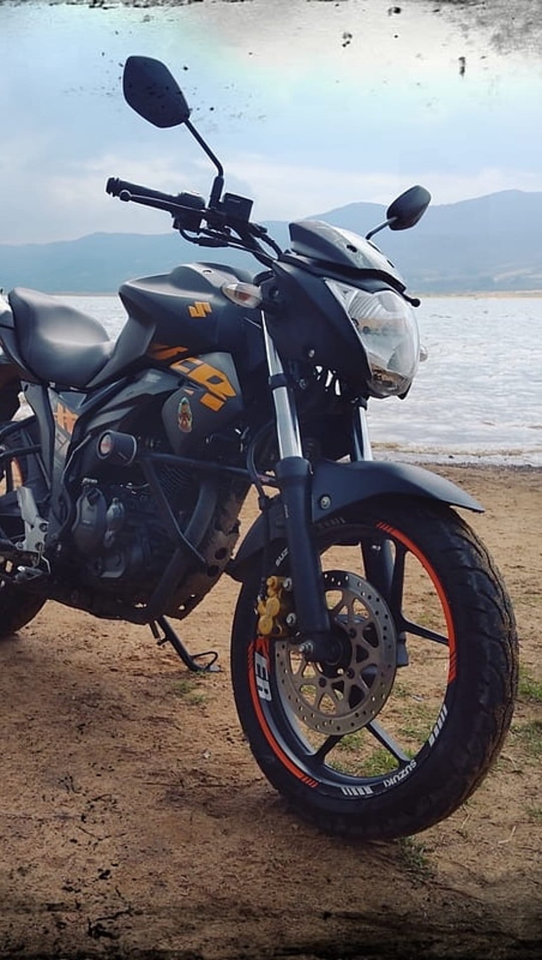 Suzuki adventure motorcycle with orange rims parked on a sandy beach near a calm mountain lake under cloudy skies.
