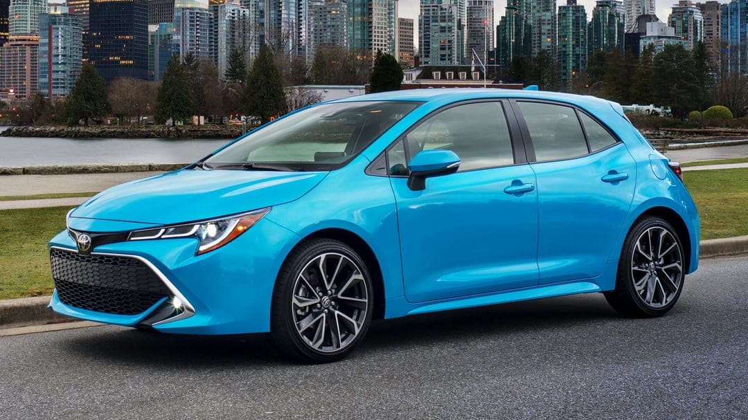 Blue Toyota Corolla Hatchback parked on a waterfront road with a towering glass city skyline in the background.