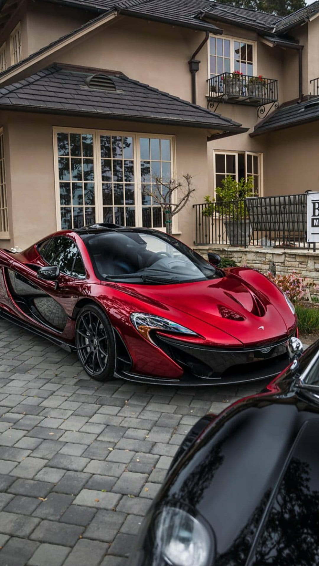 Red McLaren P1 hypercar parked on a gray cobblestone driveway in front of a modern home under bright sunlight.