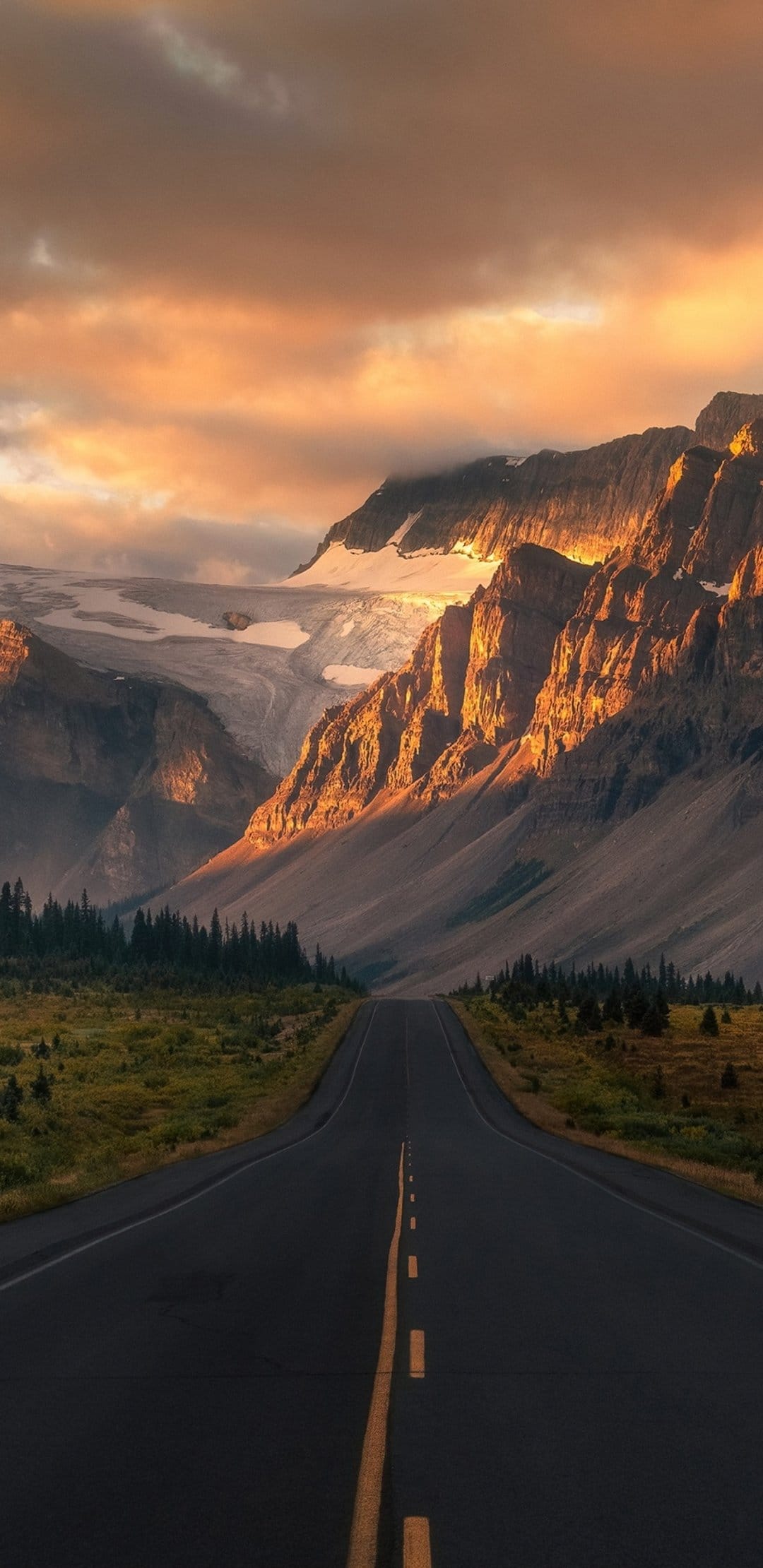Asphalt road stretching toward snow-capped mountains under a glowing golden sky with dramatic sunset clouds.