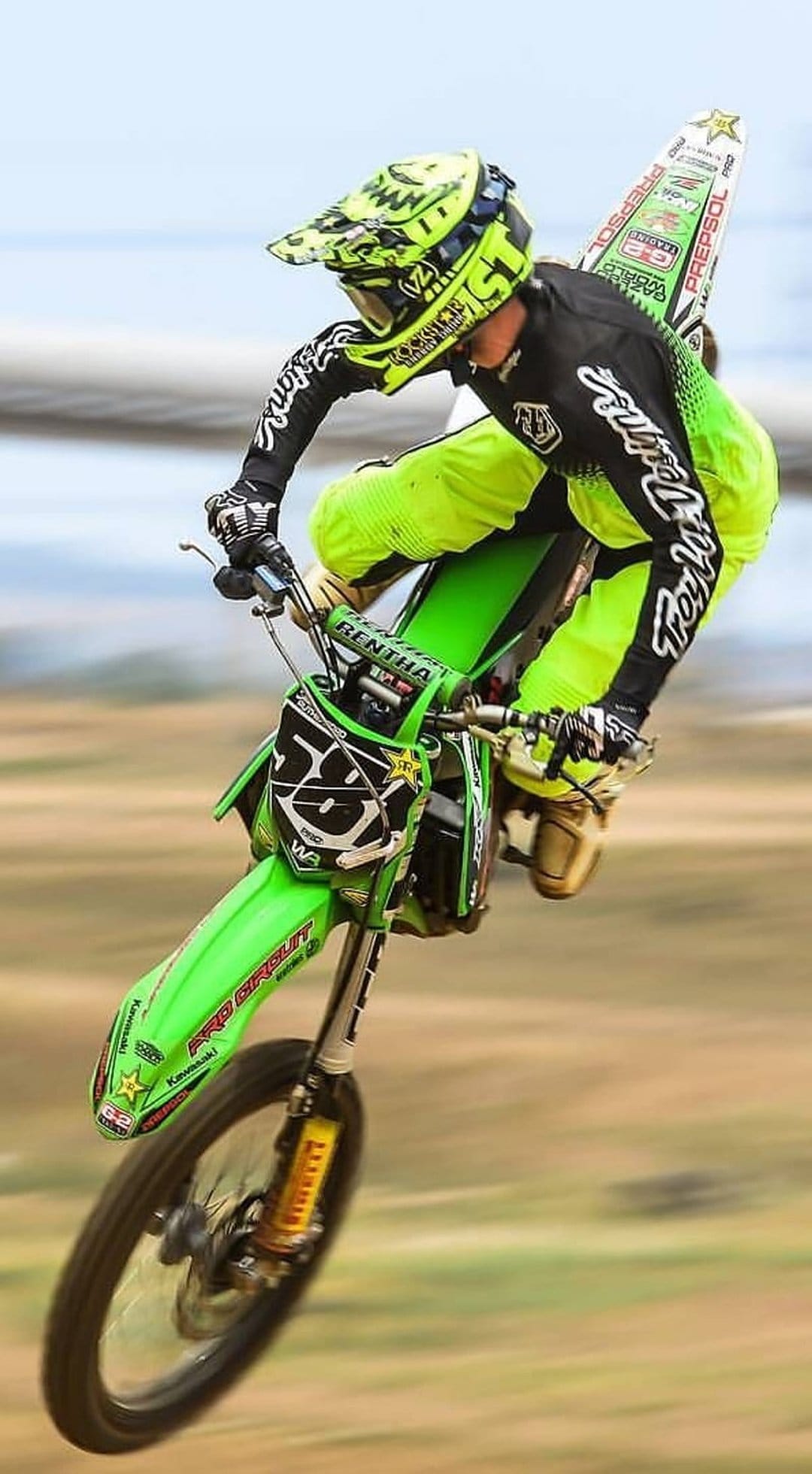 Motocross rider in neon green gear jumps a dirt bike mid-air with a yellow helmet against a blurred background.