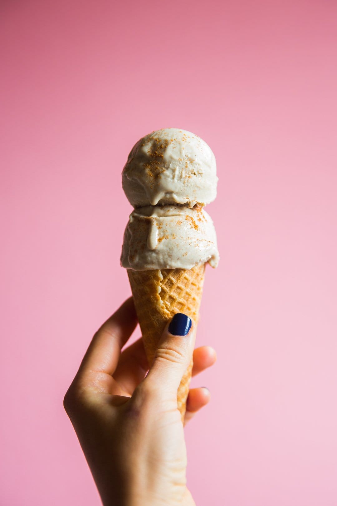 Hand with blue nails holds a double scoop waffle cone dusted with spice against a solid pink background.