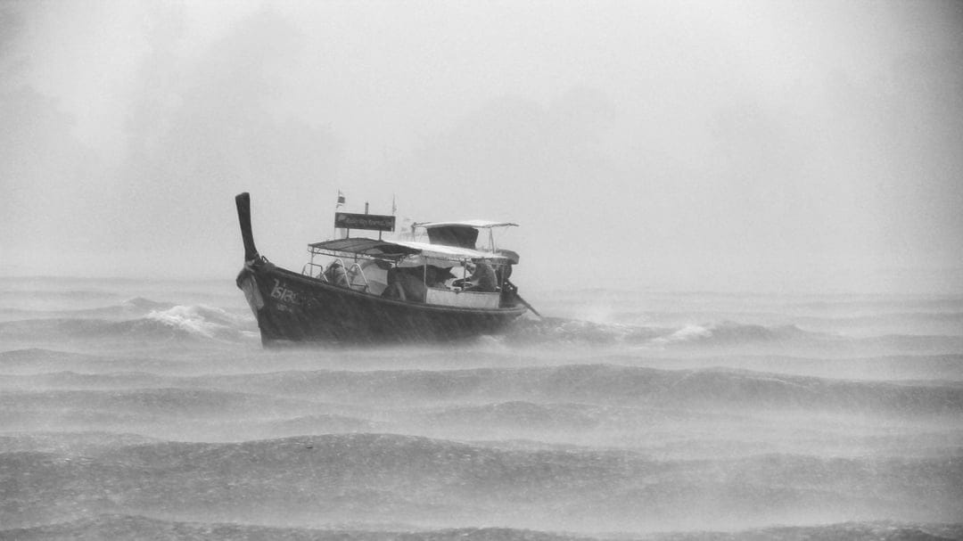 Traditional longtail boat tilting through massive crashing waves and sea spray in a dark monochrome storm.