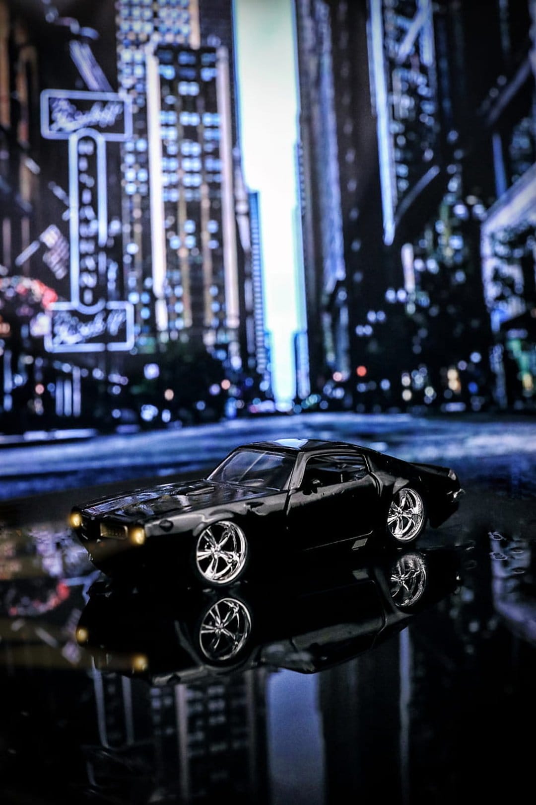 Black muscle car miniature on a wet night street with city light reflections and blurred urban skyscrapers.