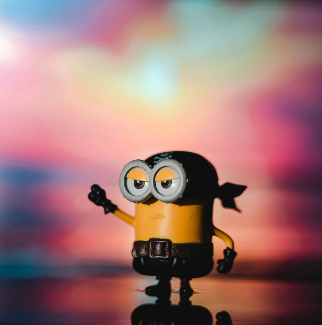 Yellow Minion toy wearing a black pirate bandana and goggles stands on a reflective surface with a pastel backdrop.