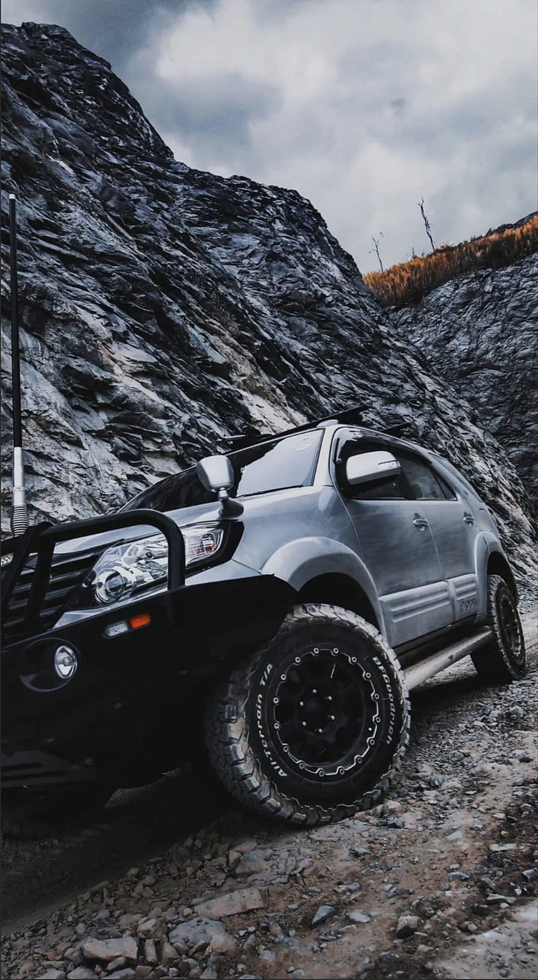 Silver SUV with a bull bar and off-road tires navigating a steep, rocky mountain trail under a cloudy sky.