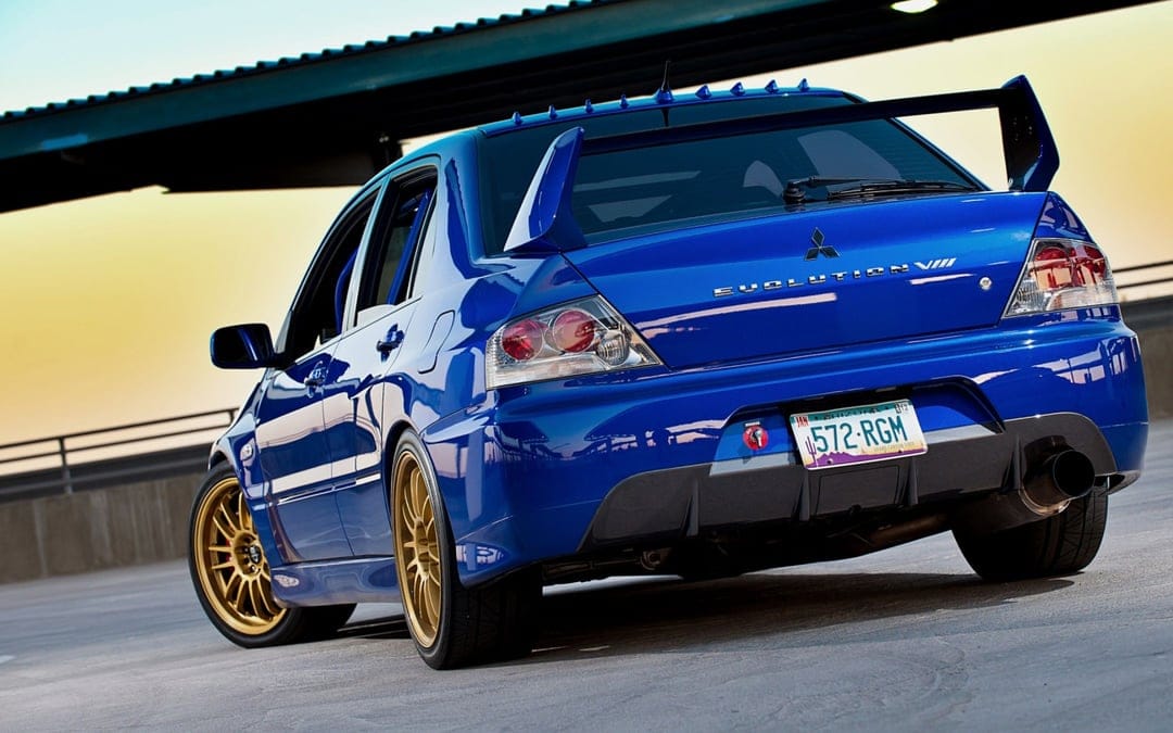 Blue Mitsubishi Lancer Evolution VIII with gold wheels and large spoiler parked on a rooftop during sunset.